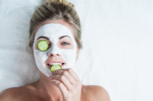 young woman with facial mask, biting on a cucumber