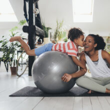 Latin woman sitting in a lotus pose on a mat, her little daughter on a pilates ball next to her, kissing her.