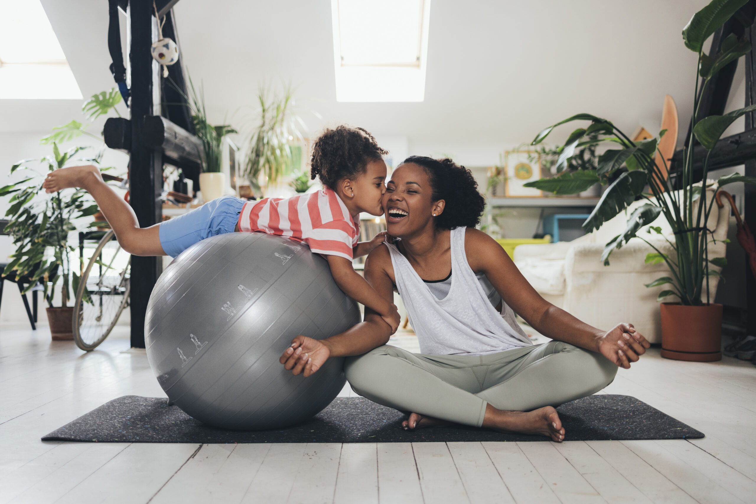Latin woman sitting in a lotus pose on a mat, her little daughter on a pilates ball next to her, kissing her.