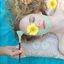 A woman with a flower over one eye gets painted with a blue facial cream.