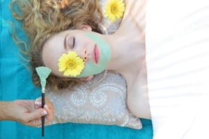 A woman with a flower over one eye gets painted with a blue facial cream.