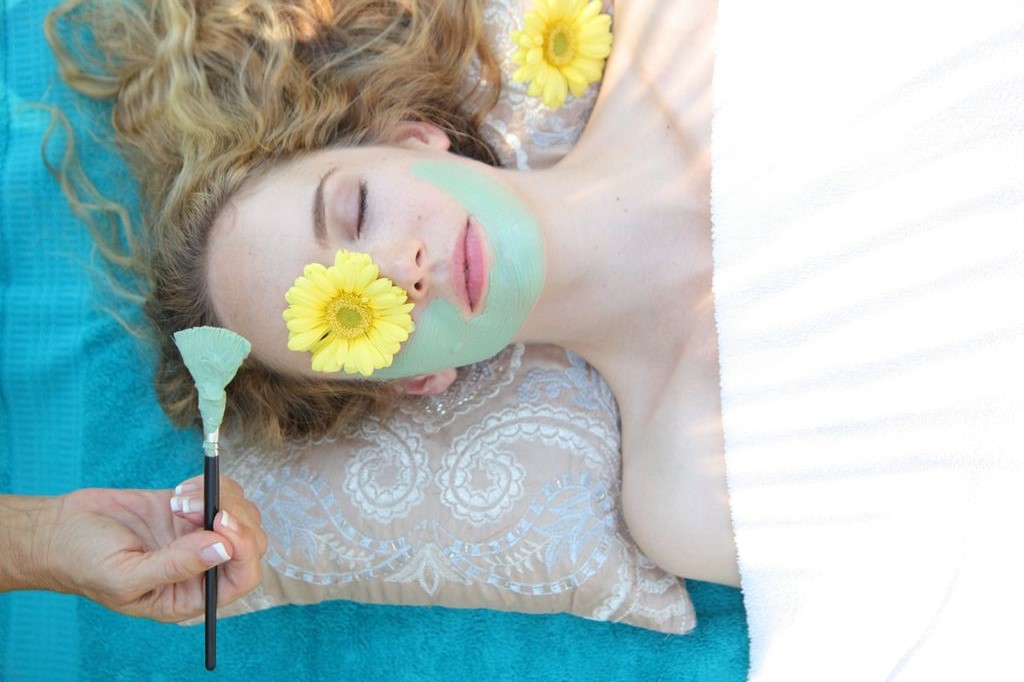 A woman with a flower over one eye gets painted with a blue facial cream.
